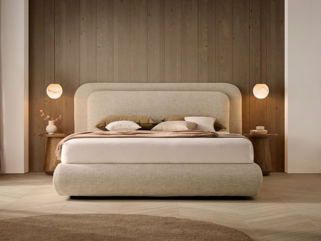 Neutral-toned bedroom with a large upholstered bed against a wood-paneled accent wall, matching nightstands, and warm round lamps.