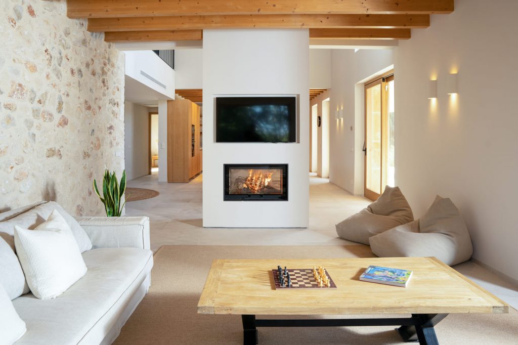 Living room with a central white column containing a fireplace under a flat-screen TV, flanked by a stone wall and exposed wooden beams.