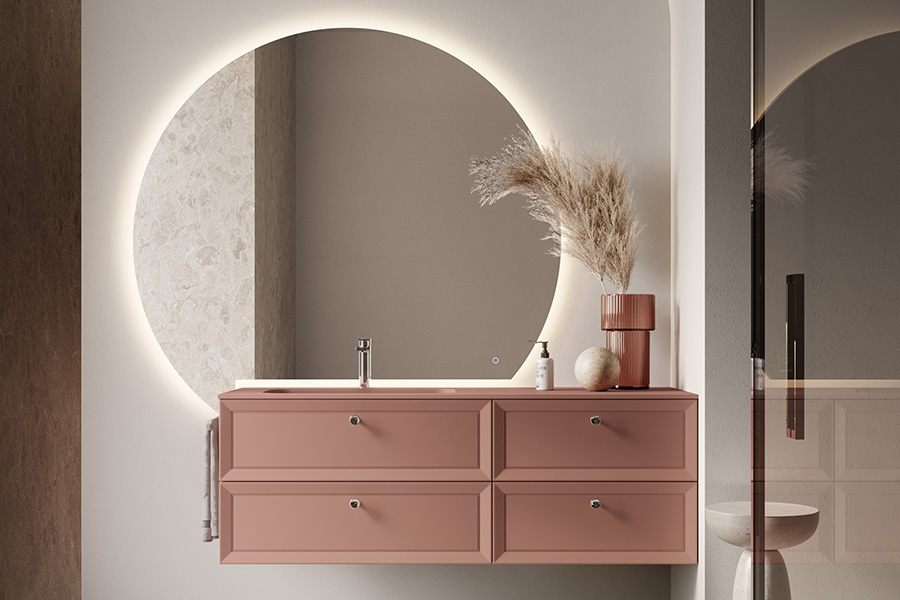 Peach-colored double-drawer bathroom vanity with a round backlit mirror, faucet, and pampas grass vase on top.