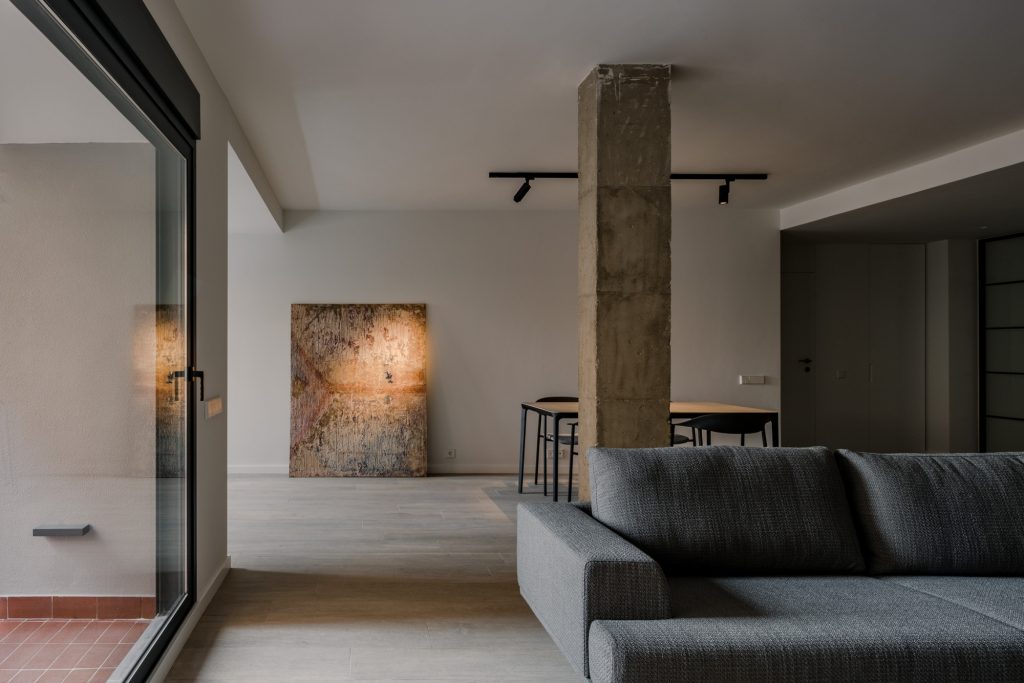 Open-plan living area with a concrete pillar, grey sectional sofa, and a dining table under track lighting.